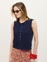 A woman in dark sunglasses and the Taylor Vest In Cotton, a sleeveless navy piece with shell buttons, stands against a white background. She has light brown hair, a colorful floral skirt, and a red scrunchie on her wrist.