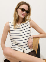 A woman wears the Taylor Striped Vest in cotton—sleeveless, white with black horizontal stripes—paired with sunglasses, sitting cross-legged on a chair against a plain white background.
