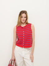 A woman with light brown hair wears the Taylor Striped Vest In Cotton— a sleeveless red vest with blue stripes—paired with white pants and holding a red woven handbag, standing against a plain white background.