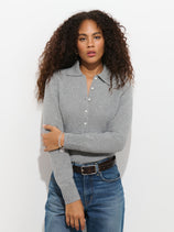 A person with curly hair models the Park Polo in Washed Cashmere, paired with jeans. They stand against a plain white background with one arm crossed and a neutral facial expression.