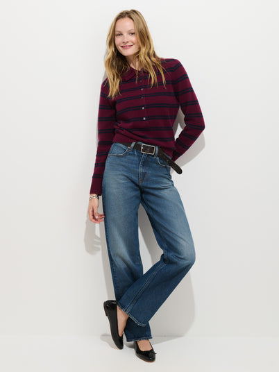 A woman stands against a white wall, smiling. She wears the Park Striped Polo in Washed Cashmere—an extra-soft maroon and navy striped long-sleeve sweater—paired with blue jeans, a black belt, and black flats. Her hair is loose and wavy.