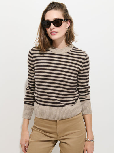 A woman stands against a plain white background, wearing the Marais Striped Sweater in Washed Cashmere—a luxurious black and beige striped piece crafted from 100% cashmere for exceptional softness. She pairs the sweater with tan pants and dark sunglasses, one hand resting in her pocket and a slight smile on her face.