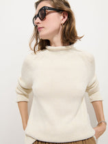 A woman with light brown hair wears black sunglasses, gold hoop earrings, and the Eastport Rollneck In Cotton—a cream-colored fisherman’s sweater made from 100% cotton. With her sleeves rolled up and hands behind her back, she looks to her left against a plain white background.