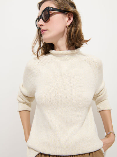 A woman with light brown hair wears black sunglasses, gold hoop earrings, and the Eastport Rollneck In Cotton—a cream-colored fisherman’s sweater made from 100% cotton. With her sleeves rolled up and hands behind her back, she looks to her left against a plain white background.