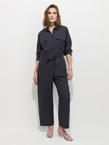 A woman stands against a white background, wearing the Delancey Jumpsuit in Lightweight Twill—a relaxed fit, dark navy cotton one-piece featuring long sleeves, chest pockets, and a drawstring waist—paired effortlessly with strappy sandals.
