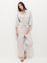 A woman stands against a white background wearing the Delancey Jumpsuit in Lightweight Twill in light gray, featuring a relaxed fit and drawstring waist, paired with tan sandals, a colorful neck scarf, and a wristwatch.
