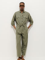 A person stands against a plain white background, hands in pockets, wearing the Delancey Jumpsuit in lightweight twill—an olive green relaxed-fit piece with rolled-up sleeves—paired with dark sunglasses.