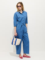 A woman in the Delancey Jumpsuit in Lightweight Twill, wearing pink sunglasses and shoes, stands against a white background holding a beige tote bag with blue handles and a red letter E.