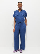 A person stands against a white background wearing the Delancey Short Sleeve Jumpsuit in Linen, garment-dyed blue with a drawstring waist and short sleeves, hands in pockets, paired with light purple shoes for an effortlessly cool summer look.