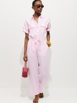 A woman in sunglasses stands against a white background wearing the Delancey Short Sleeve Jumpsuit in Linen—a light pink, garment-dyed design with a drawstring waist—paired with a red woven handbag and heeled sandals for the perfect summer look.