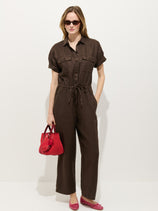 A woman stands against a white background wearing the Delancey Short Sleeve Jumpsuit in linen, styled with a red handbag, red flats, and sunglasses—an effortlessly chic summer look.
