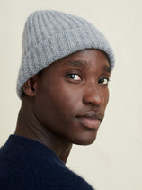 A person models the Cashmere Beanie in light gray, paired with a dark blue sweater, as they look over their shoulder toward the camera against a plain light background.